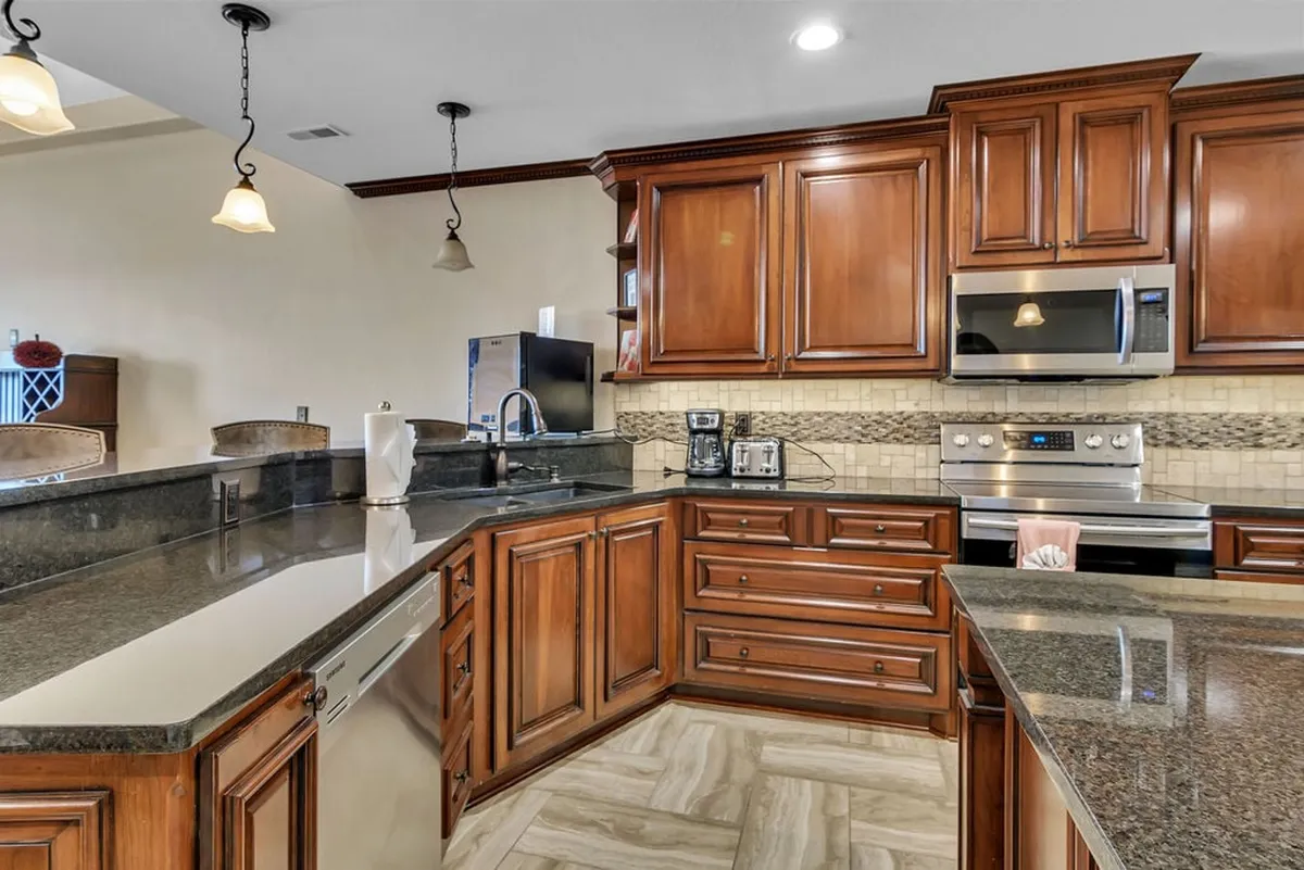 A spacious kitchen with warm wooden cabinets, gleaming granite countertops, stainless steel appliances, and a central kitchen island perfect for prepping and gathering. The space is bathed in inviting light, with ample workspace and seating designed for both meal preparation and socializing.