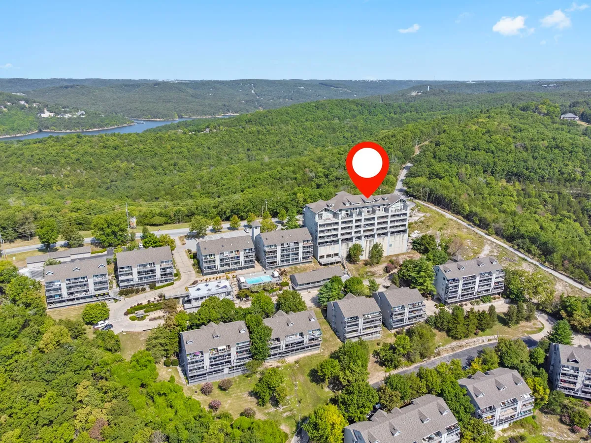 This wider exterior view captures the main building surrounded by lush greenery, a sparkling swimming pool, and a spacious parking area. The scene hints at both natural beauty and resort amenities, with the pool area inviting guests to unwind and take in the sunset after a day by the lake or in town.