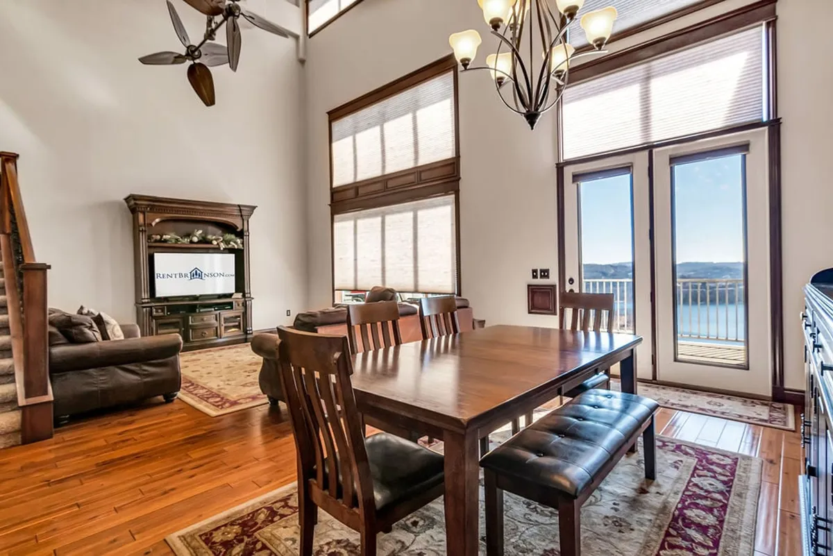 A bright, open-concept living and dining area featuring a dining table set beside large windows that offer unobstructed views of the lake. The space includes a chandelier above the table and a comfortable sofa in the living area, all surrounded by rich wood finishes and warm, inviting decor.