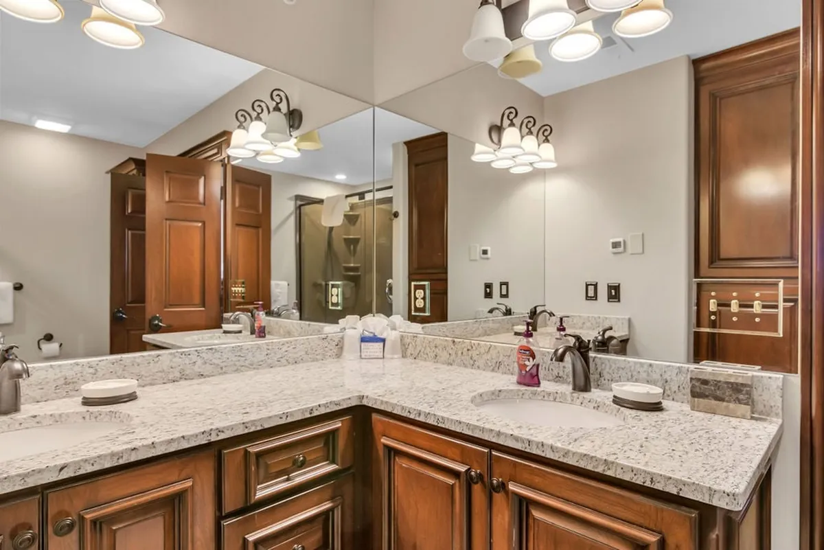 A sophisticated bathroom scene with a granite countertop, dual sinks, wood cabinetry, ample lighting, and a large mirrored wall. The overall look is modern and spa-like, offering both luxury and practicality.