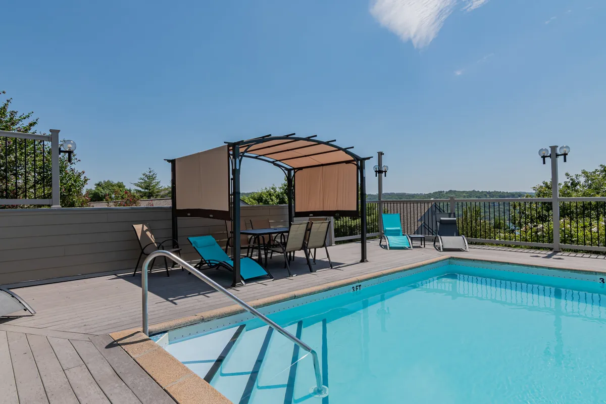 A sparkling swimming pool is framed by a canopy with a seating area, lounge chairs, and a scenic landscape in the background. The setting is ideal for leisure, sunbathing, and casual socializing in a resort-style environment.