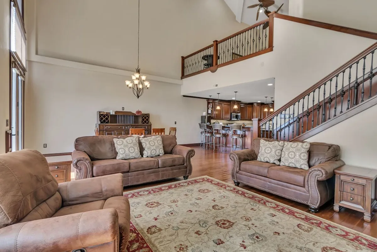 The image shows a warm and inviting living area with plush, comfortable sofas arranged around a stylish coffee table atop a decorative rug. A grand chandelier provides elegant lighting, and a staircase hints at the spaciousness of the penthouse layout, creating a perfect space for family gatherings or relaxing evenings.